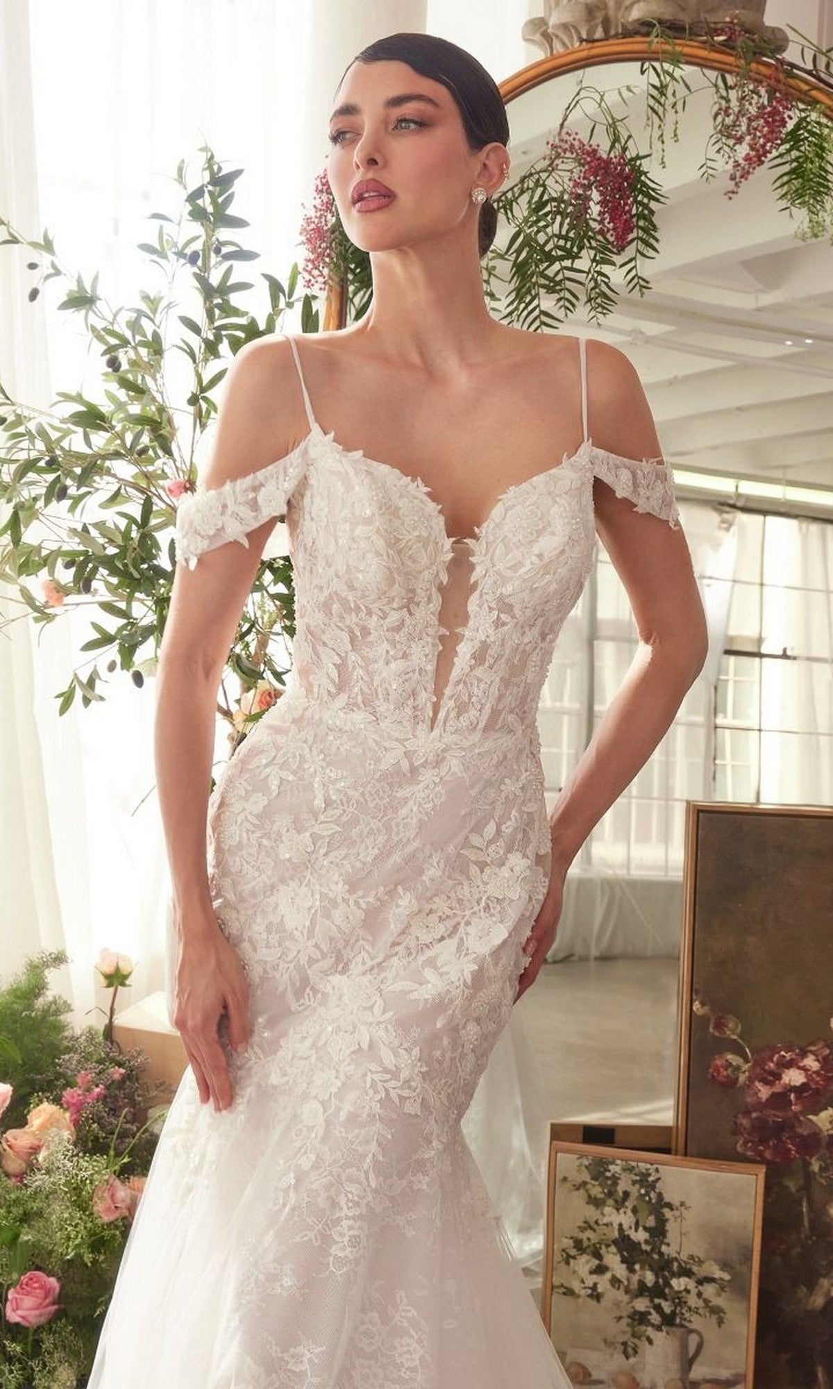 A woman in the Andrea and Leo WL051 long wedding dress with off-shoulder lace applique stands indoors by floral arrangements and a vintage mirror.